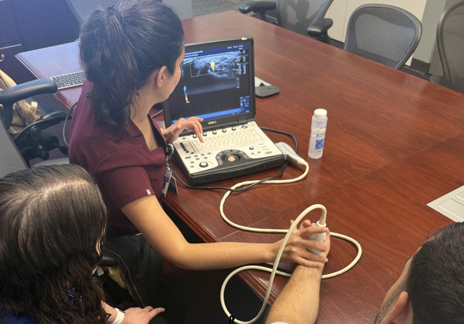 Participants gather around a portable ultrasound machine during a hands-on teaching session in a conference room. One person operates the ultrasound probe on another participant's wrist while others observe the screen.