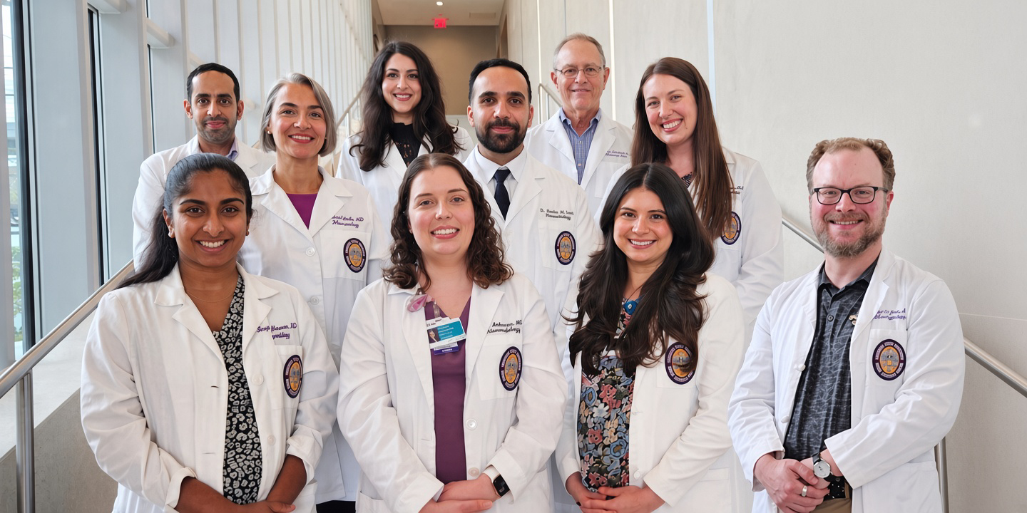 Group portrait of ten healthcare professionals wearing white lab coats, standing on a staircase in a bright, modern building with large windows. The individuals are arranged in three rows and facing the camera.