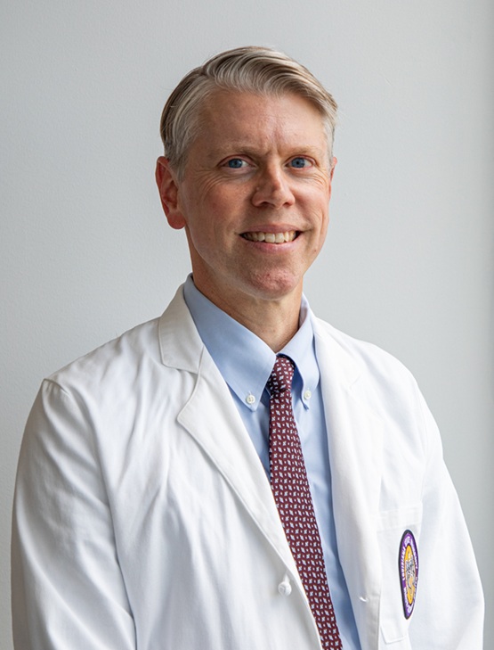 Professional headshot of a person wearing a white medical coat over a light blue dress shirt and patterned burgundy tie. The portrait is framed from the chest up against a light gray background. The subject faces slightly to one side while looking toward the camera. Soft natural lighting and a neutral backdrop create a clean, professional appearance suitable for a healthcare or academic profile.