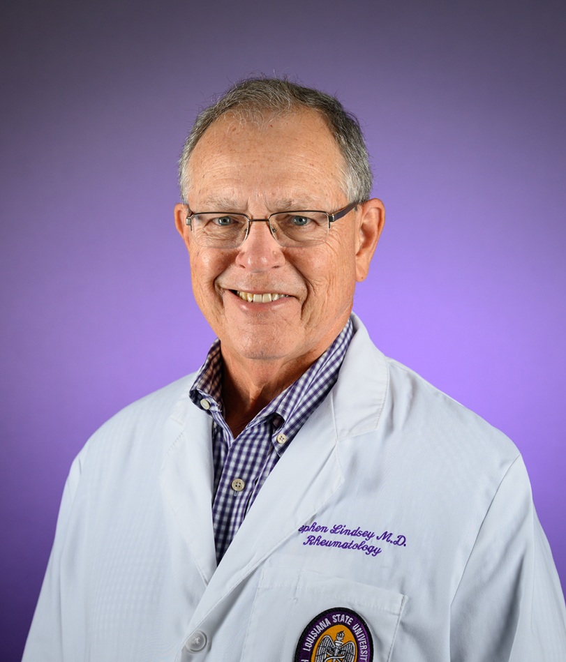 Portrait of a person wearing a white lab coat and glasses, photographed from the chest up against a purple gradient background; the lab coat has embroidered text and an institutional patch on the chest, with a checkered shirt visible underneath.