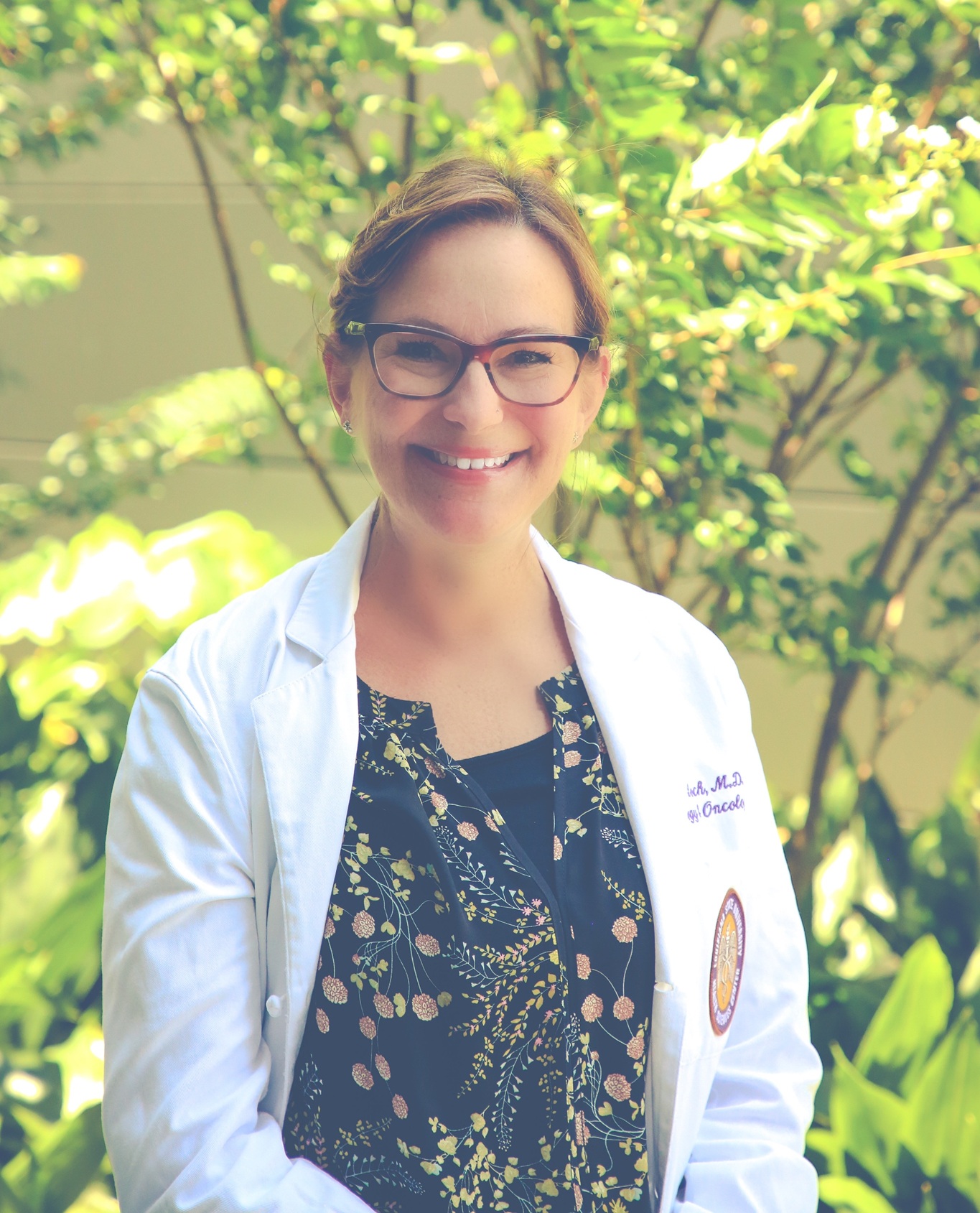 Portrait of a person wearing glasses and a white medical coat over a dark floral-patterned top, standing outdoors in front of leafy green plants. The person is facing the camera. The background is softly blurred with sunlit foliage, creating a bright natural setting. An embroidered name and a circular emblem are visible on the coat, though the text is not fully legible.