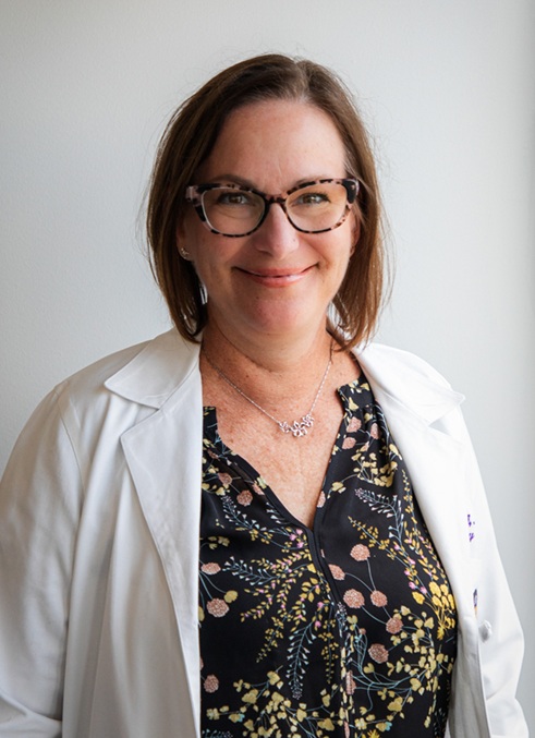 Professional headshot of a person wearing a white medical coat over a dark floral-patterned blouse and eyeglasses. The portrait is framed from the waist up against a light gray background. The subject faces the camera in a neutral pose, with soft, even lighting creating a clean and professional appearance suitable for a healthcare or academic profile.