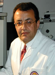 Professional portrait of a person in a white medical coat and red tie, seated in a clinical setting with medical equipment in the background.