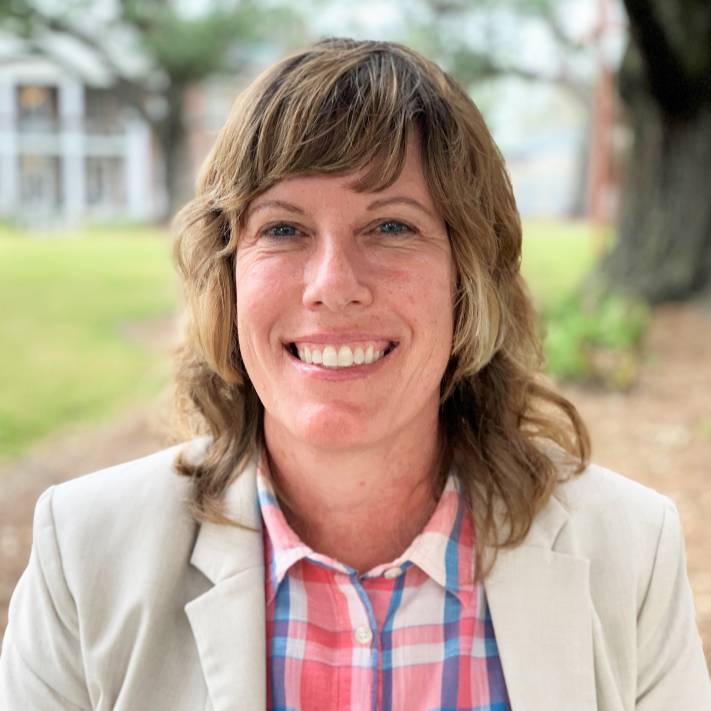 Outdoor professional portrait of a person seated in a landscaped campus setting. The person has shoulder-length wavy hair and is wearing a light beige blazer over a pink, blue, and white plaid button-down shirt. The person is facing the camera, with large trees, grass, and a building softly blurred in the background. Natural daylight and a shallow depth of field create a polished headshot appearance.
