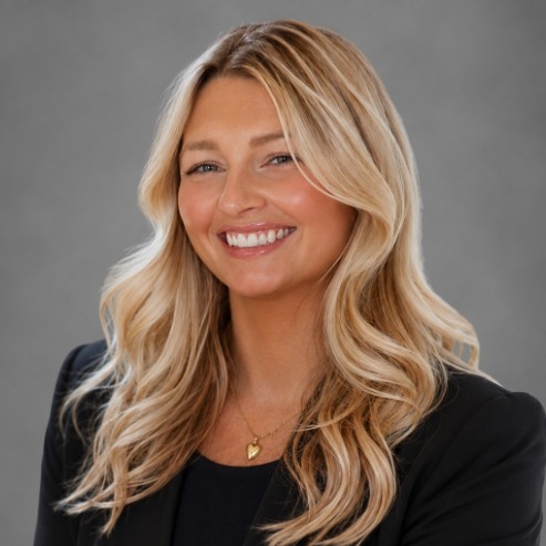 Professional studio portrait of a person with long, wavy blonde hair wearing a black blazer over a dark top and a small pendant necklace. The person is seated facing the camera against a neutral gray background. Soft, even lighting creates a polished headshot with the upper body visible.
