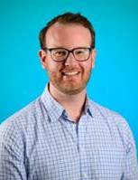 Professional head-and-shoulders portrait of a person wearing glasses and a light blue checkered button-down shirt, facing the camera against a bright blue studio background. The image is well-lit with a clean, simple composition.