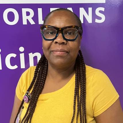 Portrait of a person standing indoors in front of a purple wall with partially visible text, including “Health” and “New Orleans.” The person is wearing large dark-framed glasses, a yellow short-sleeved top, and a staff badge clipped near the shoulder. Long braided hair falls over the front of the top. The image is brightly lit, with the purple and yellow colors creating a strong visual contrast.