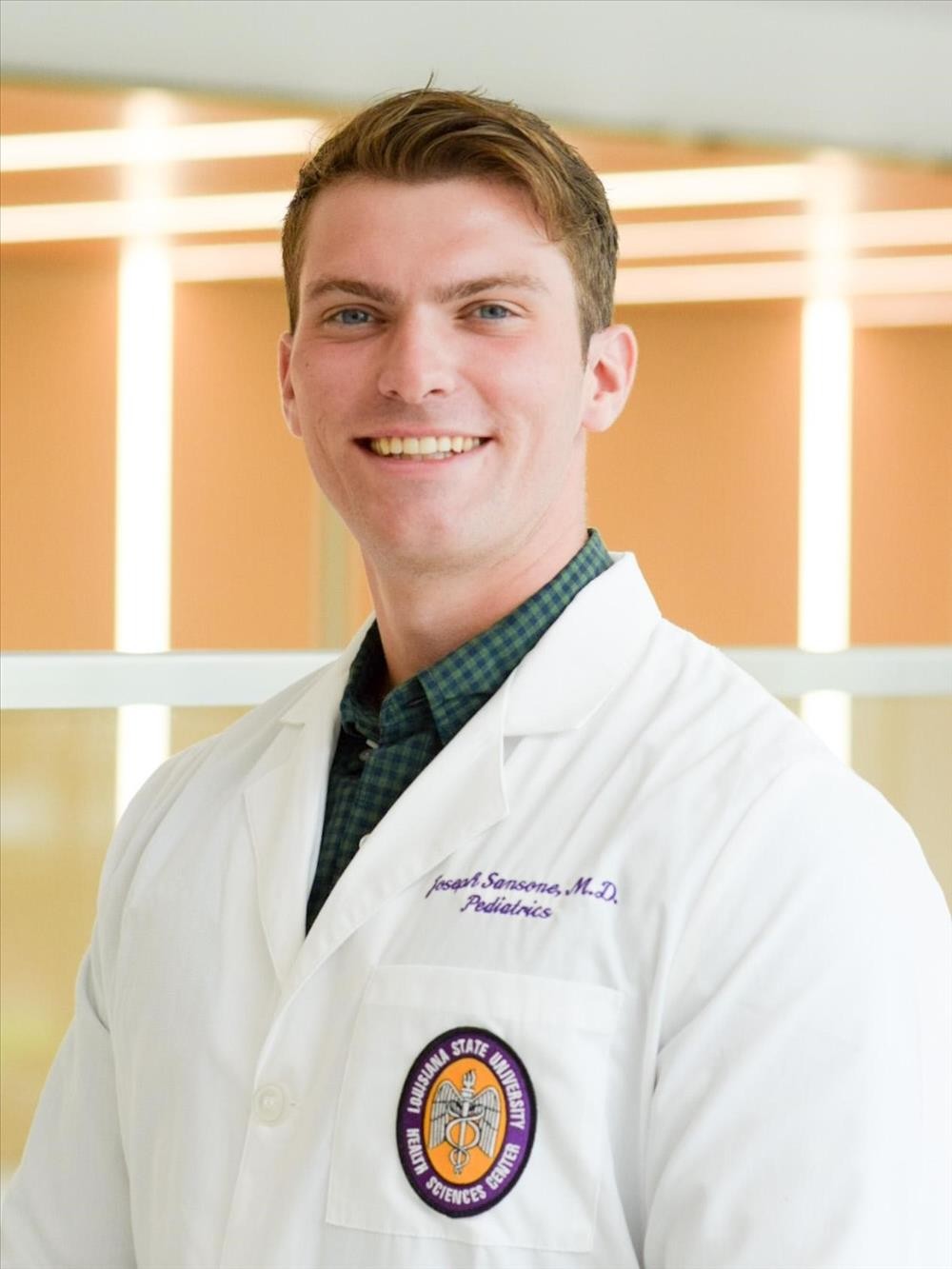 Professional headshot of an individual in a white medical coat with a university health sciences emblem, photographed in a bright indoor setting.