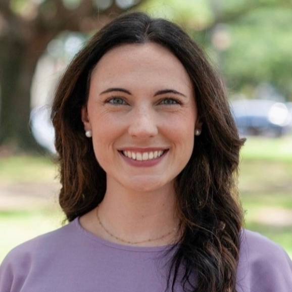 Outdoor head-and-shoulders portrait of a person with long dark hair wearing pearl stud earrings and a light purple top. The person is facing the camera in a park-like setting with large trees and greenery in the background. The background is softly blurred, with natural daylight illuminating the scene.