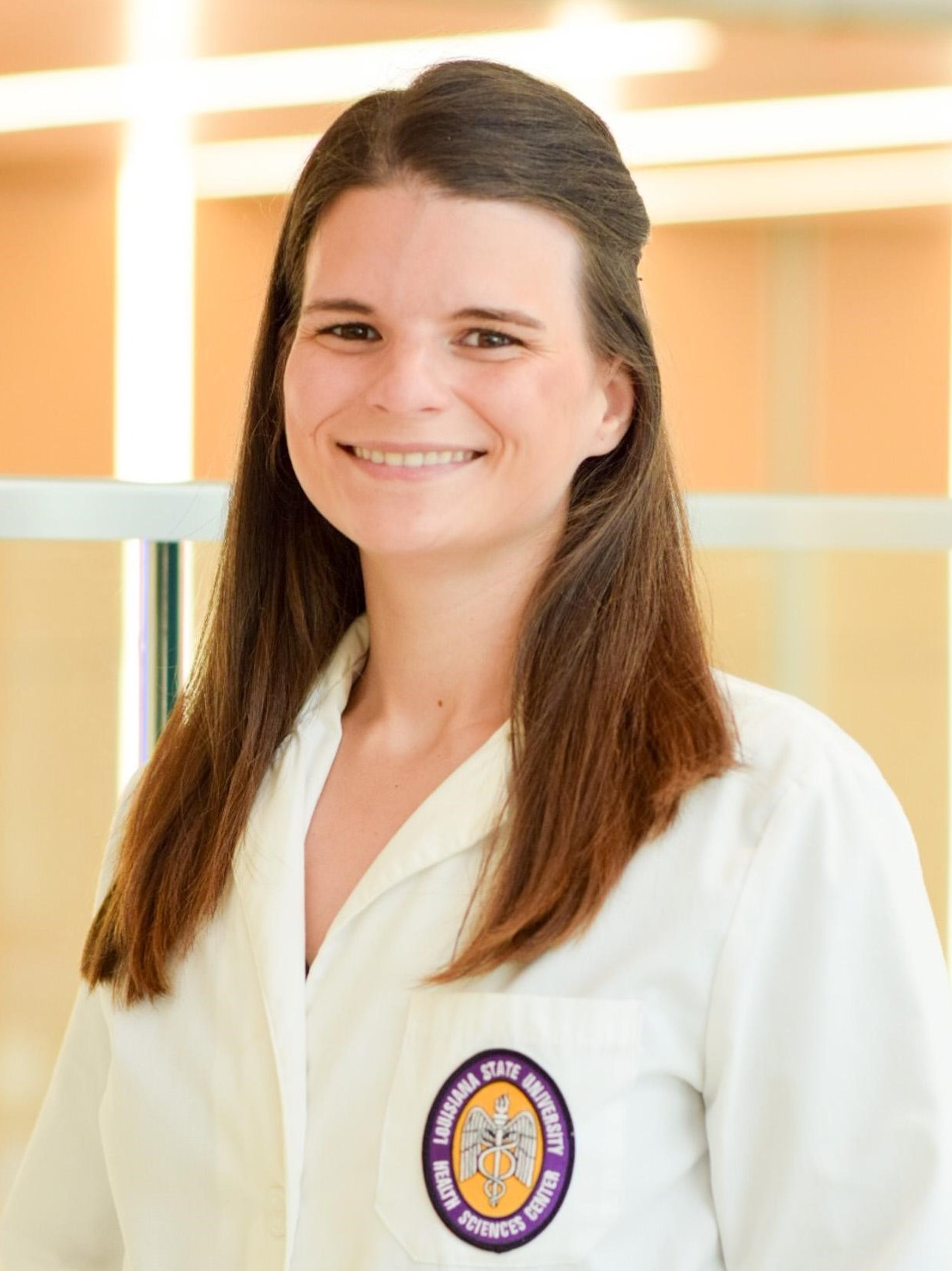 Professional headshot of a person with long dark hair wearing a white lab coat. The coat features a Louisiana State University Health Sciences Center patch on the left pocket. The person is facing the camera indoors, with large windows and warm natural light in the background.