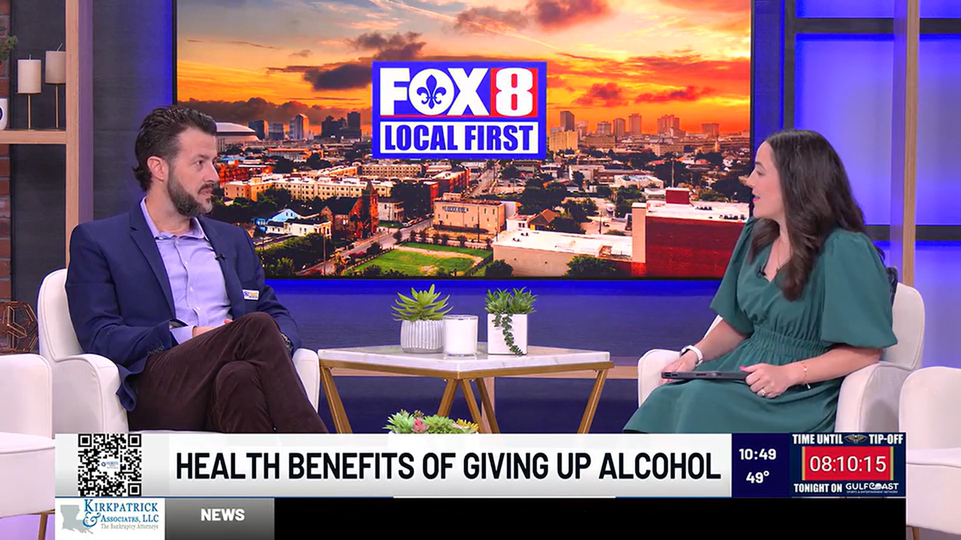 A television studio interview scene showing two individuals seated in white armchairs on either side of a small round table with plants and a mug. Behind them is a large screen displaying a bright sunset cityscape with the “FOX 8 Local First” logo centered on it. One individual on the left is seated with legs crossed, wearing a blazer, while the individual on the right holds a tablet and wears a short‑sleeved dress. At the bottom of the screen, a banner reads “HEALTH BENEFITS OF GIVING UP ALCOHOL.” A QR code appears on the left side of the banner. On the right side, a digital countdown clock shows “08:10:15,” along with the temperature reading of 49°.