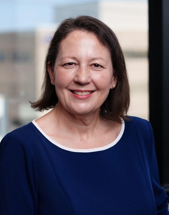 Professional headshot of a person in a blue blouse, posed in front of a blurred office background.