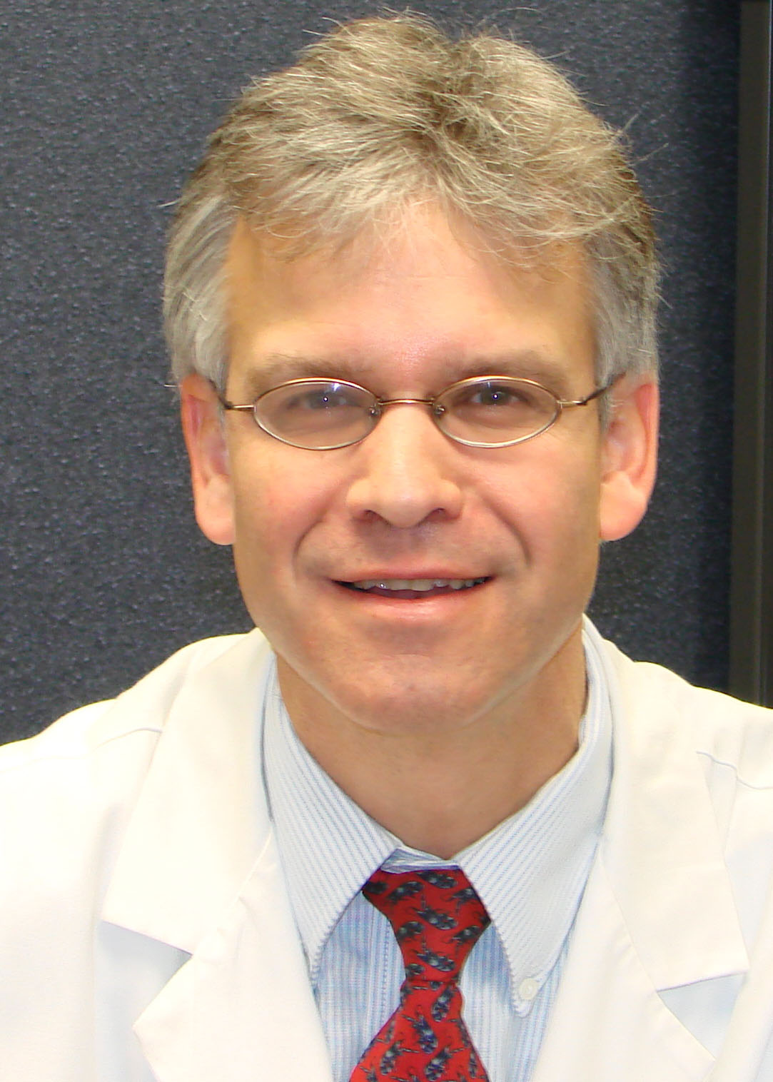 Professional headshot of a person wearing glasses, a white medical coat, and a red patterned tie against a dark background.