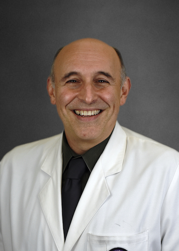 Professional headshot of a person wearing a white lab coat over a dark shirt and tie, facing the camera against a plain gray background. The person is centered in the frame, with a bald head and hair visible at the sides. The portrait is evenly lit and cropped from the chest up, in a typical corporate or medical-profile style.