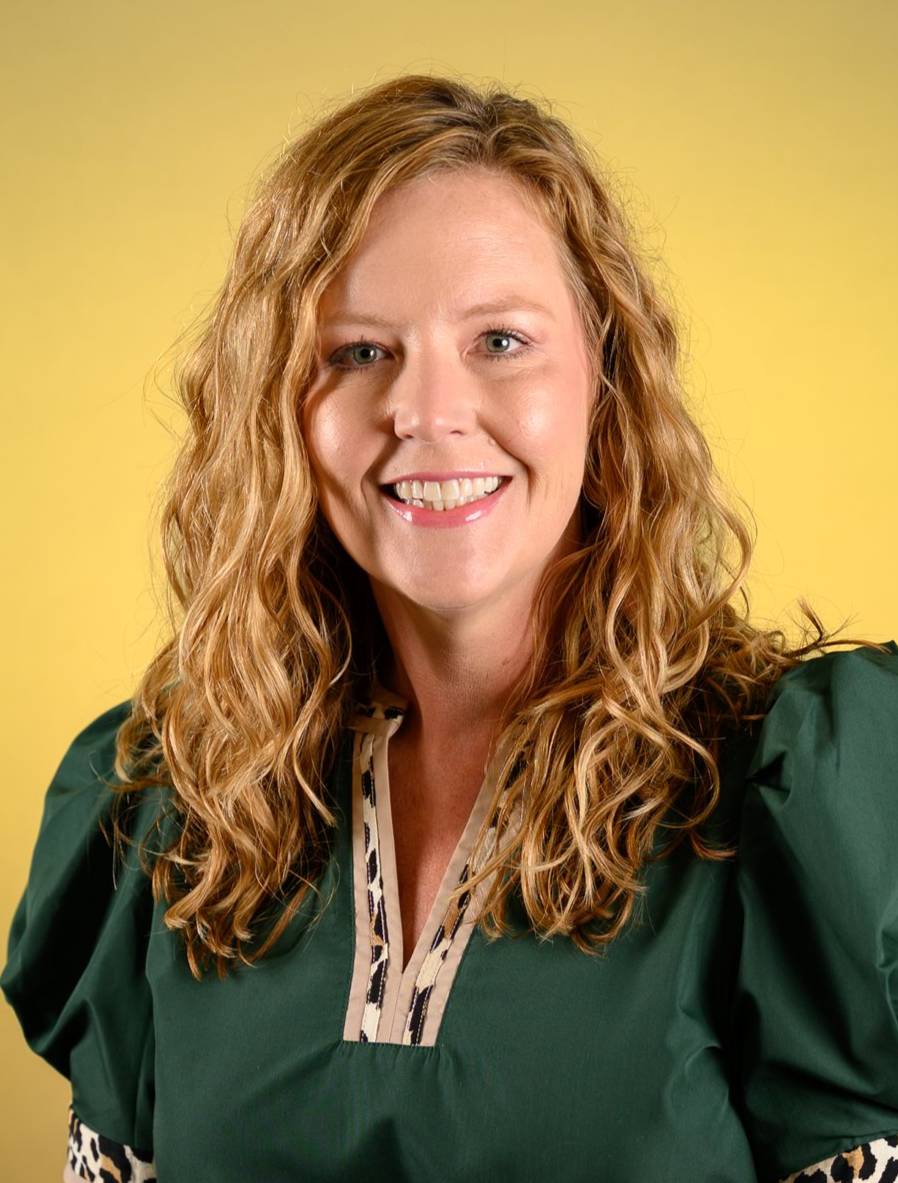 Professional headshot of a person with wavy shoulder-length hair wearing a dark green blouse, posed against a gold background.