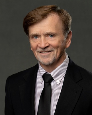 Professional headshot of a person in a dark suit, striped shirt, and black tie against a gray background.