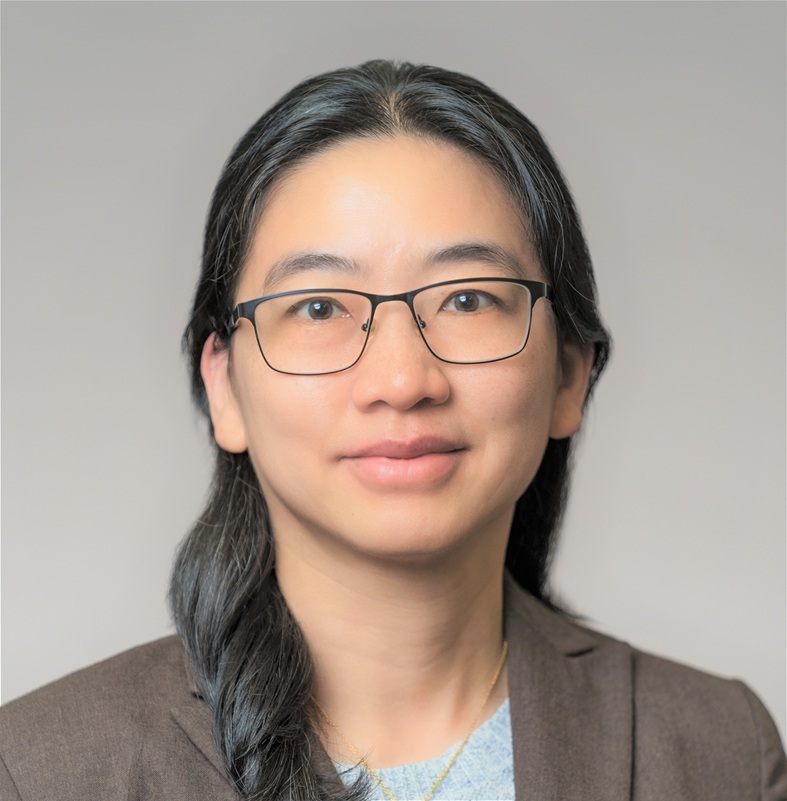 Professional head-and-shoulders portrait of a person wearing black rectangular eyeglasses, a brown blazer, and a light-colored top. The person has long dark hair worn over one shoulder and is facing the camera against a plain light gray studio background. The image is evenly lit, cropped from the shoulders up, and presented in a formal professional portrait style. Provide your feedback on BizChat