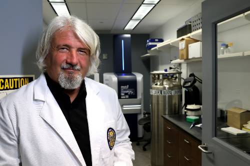Person wearing a white laboratory coat over a black shirt, standing in a research laboratory. Laboratory equipment, storage cabinets, countertops, and scientific instruments are visible in the background, including a large cylindrical tank and a workstation behind a glass door. The image is brightly lit and depicts a professional scientific research environment.