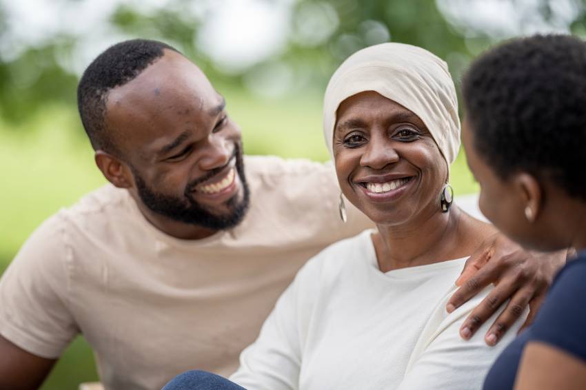 A smiling cancer patient wearing a headscarf sits outdoors with two loved ones, sharing a moment of warmth and support. The scene conveys hope, connection, and resilience during the cancer journey.