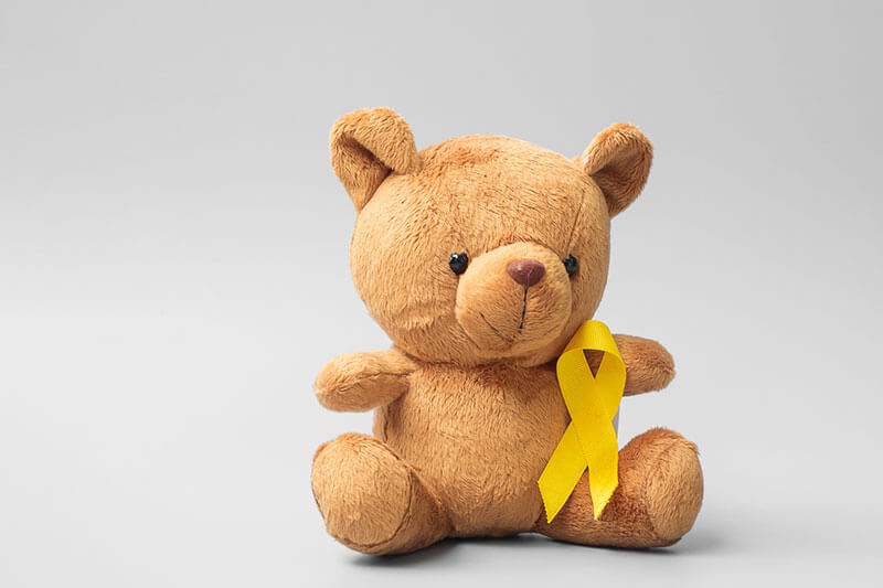 Teddy bear sitting on a light background with a gold childhood cancer awareness ribbon tied around its neck.