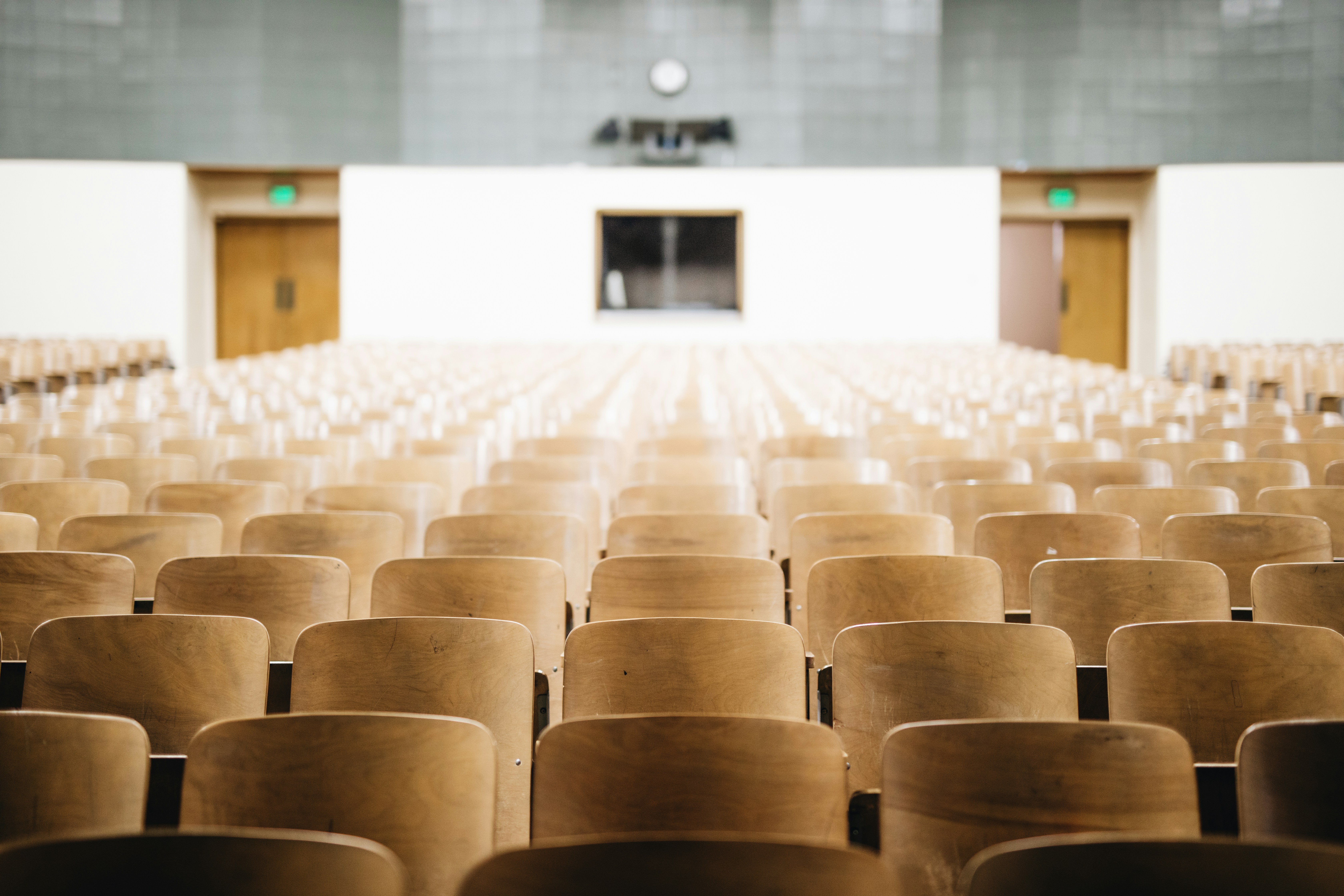 Empty lecture hall with rows of wooden chairs facing a blank stage and wall-mounted clock.
