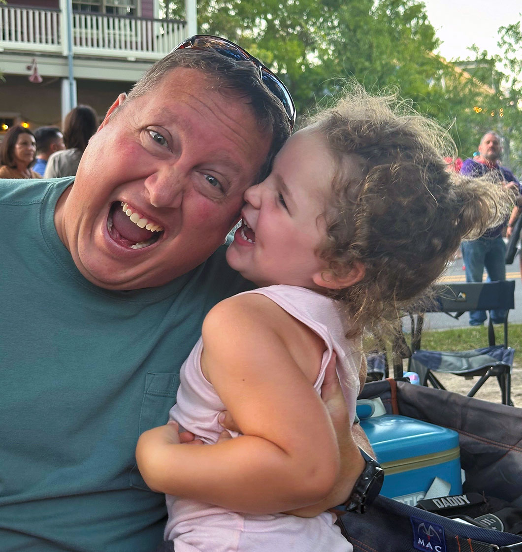 Brian, a lymphoma survivor and advocate for an NCI-designated cancer center in Louisiana, shares a joyful moment with his young daughter at an outdoor gathering, both smiling and laughing together.