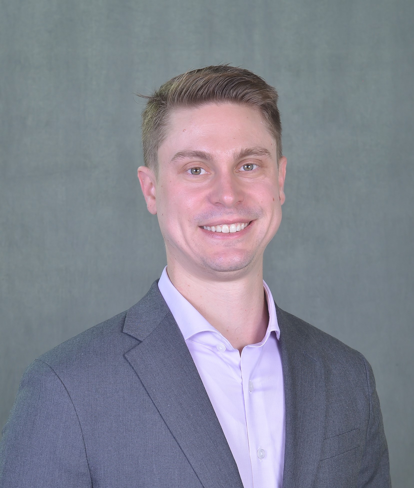 Head-and-shoulders studio portrait of a person wearing a gray suit jacket over a light-colored collared shirt, facing the camera against a plain gray background.