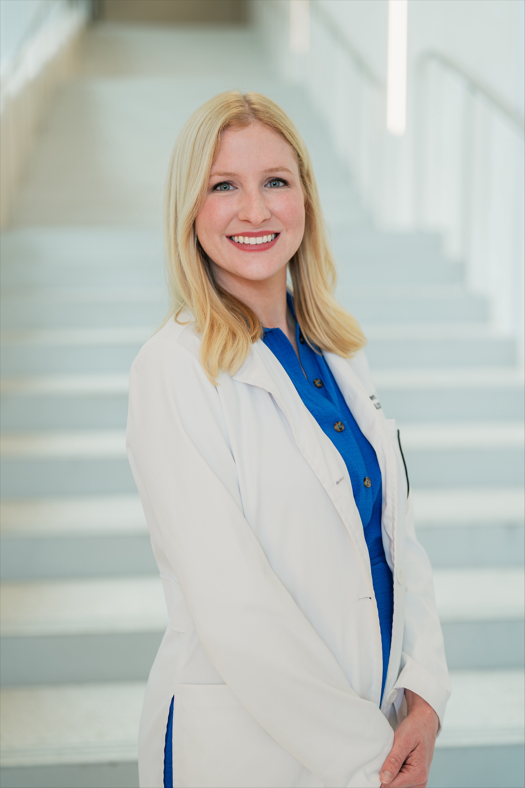 Portrait of a person with shoulder-length blond hair wearing a white lab coat over a blue button-front shirt, standing with hands clasped in front of a softly blurred staircase. The image is brightly lit with a professional indoor setting and a neutral background.