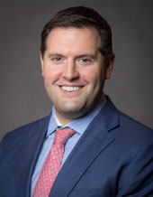Head-and-shoulders studio portrait of a person wearing a blue suit jacket, light dress shirt, and pink patterned tie, facing slightly toward the camera against a dark gray studio background.
