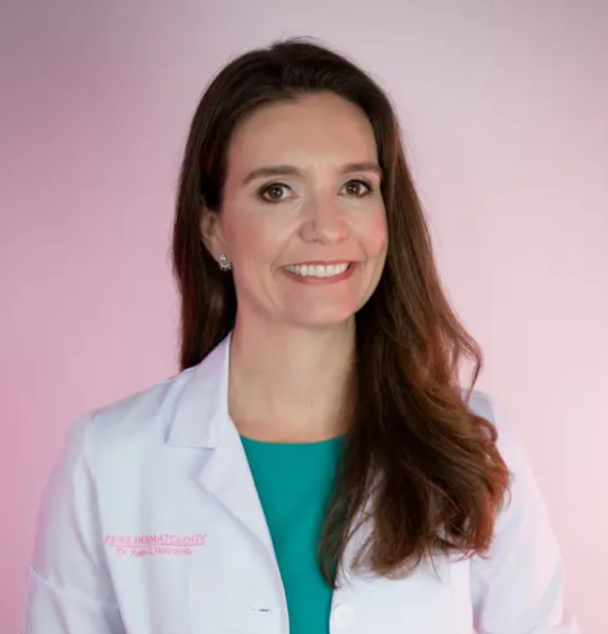Waist-up studio portrait of a person with long hair wearing a white medical-style coat over a teal dress, standing with hands in the coat pockets against a softly lit pink background. Subtle embroidered text is visible on the coat.
