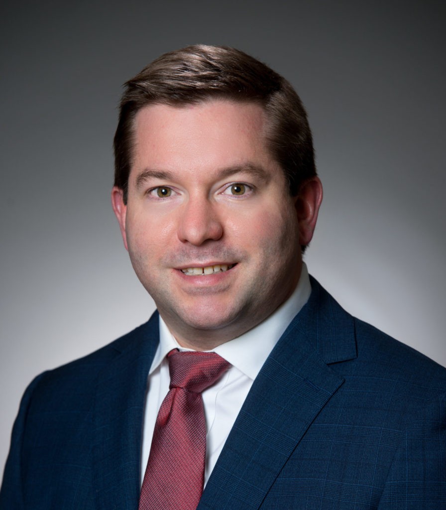 Color studio head‑and‑shoulders portrait of an adult person facing the camera against a dark gray gradient background. The person wears a dark suit jacket, a white collared shirt, and a red tie. Hair is short and neatly styled. Lighting is soft and directional, creating a sharp, professional composition.