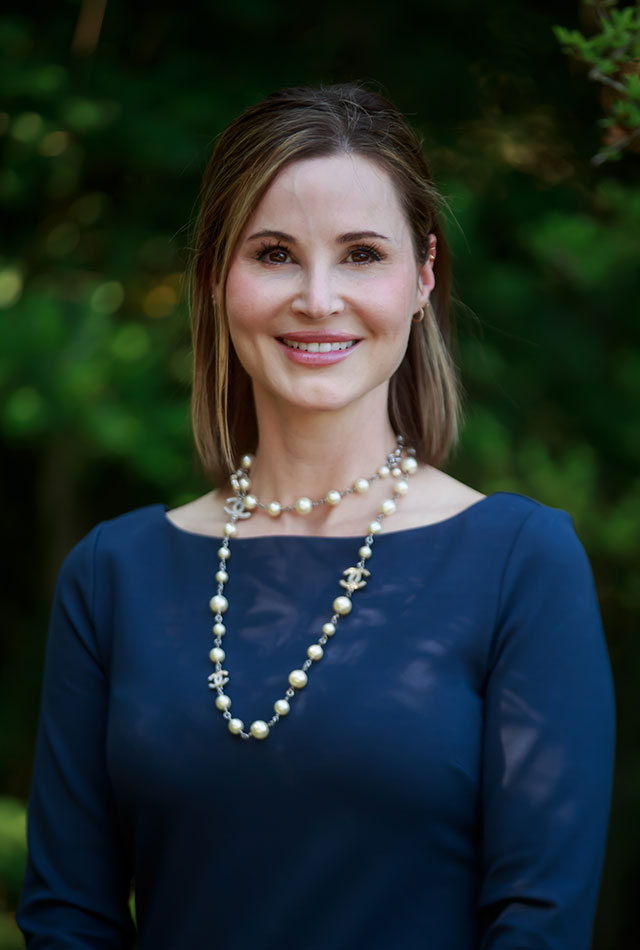 Head-and-shoulders outdoor portrait of a person wearing a navy long-sleeve top and a layered pearl necklace, standing against a softly blurred background of green foliage.