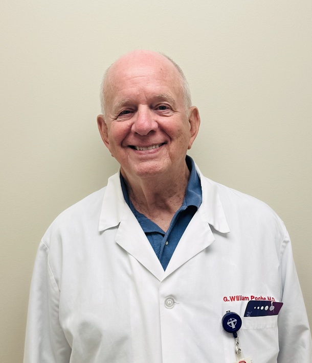A portrait of a person wearing a white medical coat over a blue collared shirt, standing against a plain light-colored wall. The person is facing forward with a slight smile. A name embroidered in red text is visible on the coat, along with an ID badge clipped near the chest pocket.