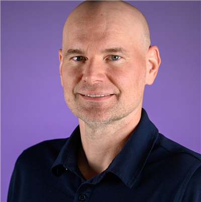Professional head-and-shoulders portrait of a person with a shaved head wearing a dark blue collared shirt. The person is facing the camera against a solid purple background, with studio lighting and a neutral, forward-facing pose.
