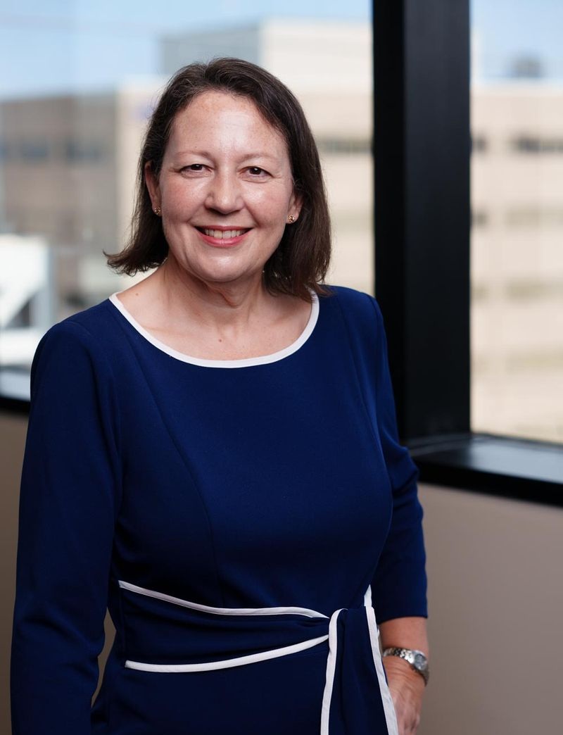 Person standing indoors near a large window, facing the camera. The person is wearing a dark blue knee-length dress with white trim at the neckline and waist tie. One hand rests at the side while the other is partially visible. A blurred office building is visible through the window in the background.