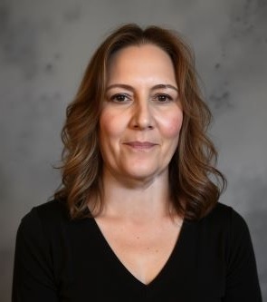 Headshot of a person with shoulder-length wavy hair wearing a black V-neck top against a gray studio background.