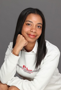 Studio portrait of a person with long, straight dark hair wearing a white sweatshirt. The person is seated with their chin resting on one hand and is facing the camera. The background is a plain gray backdrop with soft, even studio lighting. Visible text on the sweatshirt includes a red and black logo.