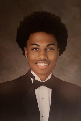 Formal studio portrait of a person with a full curly hairstyle wearing a black tuxedo jacket, white dress shirt, and black bow tie. The person is facing the camera against a mottled brown studio backdrop. Small stud earrings are visible, and the image is lit with traditional portrait-style studio lighting.