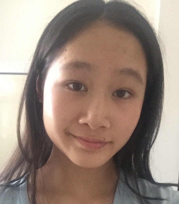 Close-up portrait of a person with long, dark hair wearing a light blue top. The person is facing the camera indoors, with a plain light-colored wall and part of a doorframe visible in the background. The image is softly lit by natural light and framed tightly around the face and shoulders.