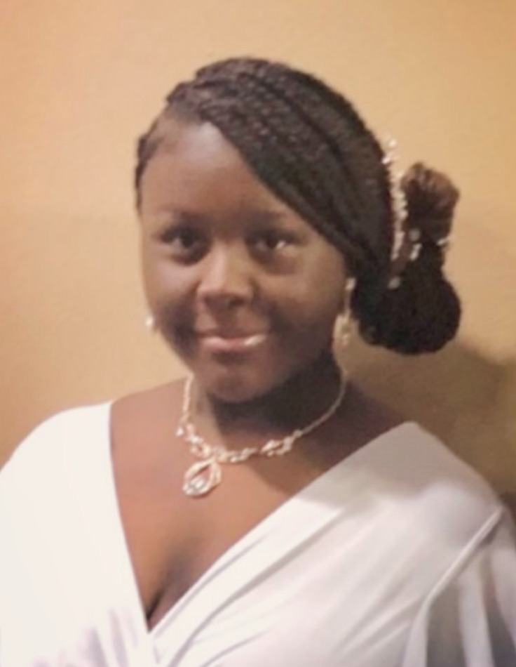 Portrait of a person with braided hair styled to one side and gathered into a low bun, wearing a white V-neck formal gown. A decorative necklace with a pendant and matching earrings are visible. The person is facing the camera against a plain beige indoor wall.