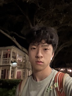 Nighttime outdoor portrait of a person with short, dark hair wearing a light-colored T-shirt and a backpack with visible shoulder straps. The person is facing the camera. In the background, a large tree and a multi-story residential-style building with balconies and illuminated windows are visible. The image is framed from the chest up and lit by ambient outdoor lighting at night.