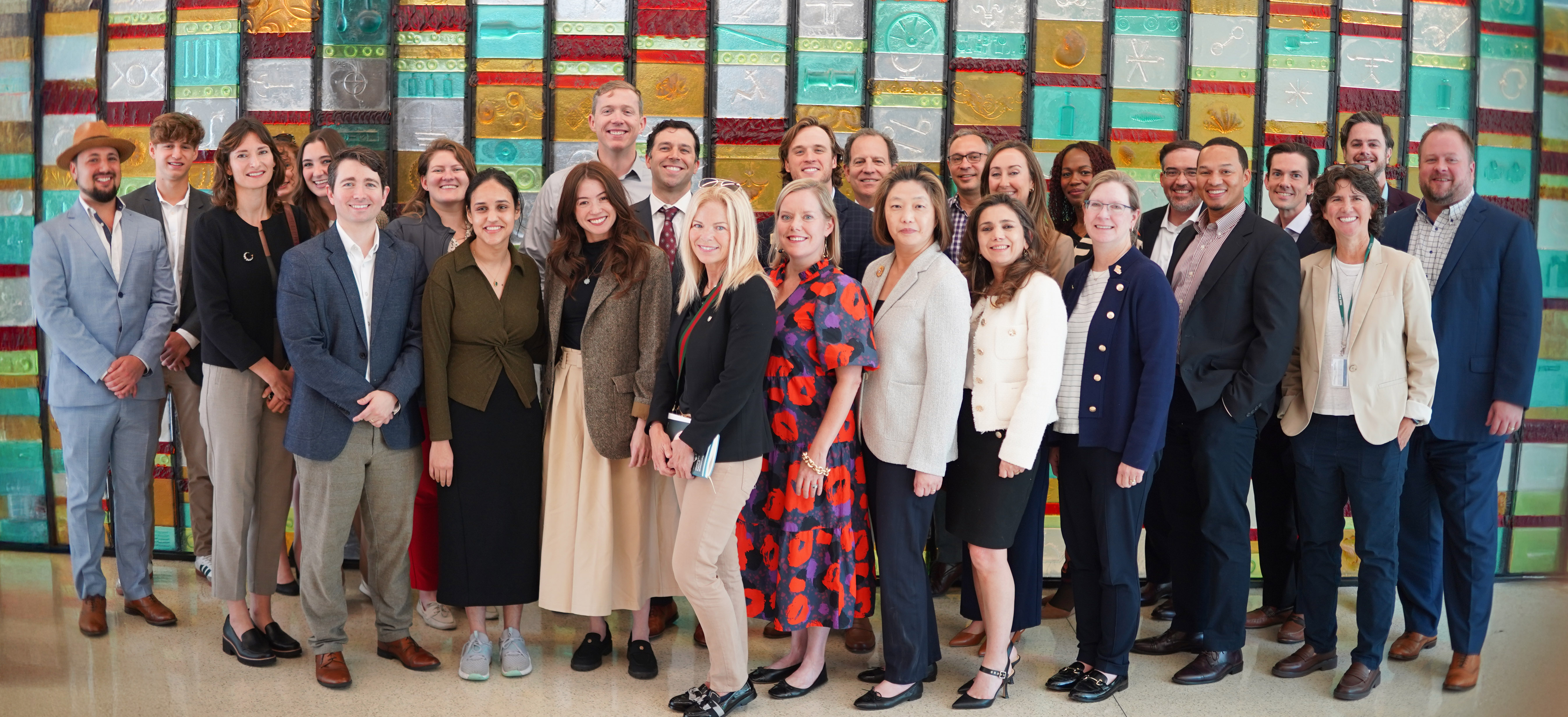 A large group of professionals stands together in a bright, modern interior, arranged in a loose semicircle as they pose for a group photo. Their attire ranges from business formal to smart casual, with suits, dresses, and blazers creating a polished, cohesive look. Behind them stretches a striking wall made of colorful rectangular panels, each embossed with unique patterns and symbols in shades of teal, red, gold, and silver. The vibrant backdrop contrasts with the group’s neutral clothing tones, giving the scene an energetic, contemporary feel. The overall impression is one of a team or cohort gathered for a formal event, workshop, or milestone moment.