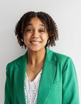 Studio head-and-shoulders portrait of a person with short, curled hair wearing a bright green blazer over a white patterned blouse. The person is facing the camera against a light gray background.
