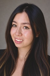 Professional head-and-shoulders portrait of a person with long, dark hair wearing a dark top. The person is facing the camera against a neutral gray studio background. The image is closely framed around the head and shoulders and lit with soft, even studio lighting.