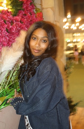 Indoor portrait of a person with long, dark hair wearing a dark denim jacket. The person is standing beside a large floral arrangement featuring bright pink flowers and tall, feathery beige stems. Warm decorative lights and an indoor walkway are visible in the softly blurred background. The image is lit with warm ambient lighting and framed from the waist up.