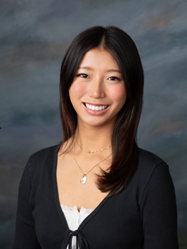 Professional head-and-shoulders studio portrait of a person with straight, shoulder-length dark hair wearing a dark cardigan over a light-colored top. The person is facing the camera against a blue-gray mottled studio background. Two delicate necklaces are visible. The image is framed from the chest up and lit with soft, even studio lighting.