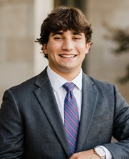 Professional outdoor portrait of a person wearing a gray business suit, light blue dress shirt, and striped tie. The person is facing the camera with one arm resting on a ledge. The image is framed from the chest up, with a softly blurred architectural background in neutral tones. Natural lighting provides clear, even illumination.