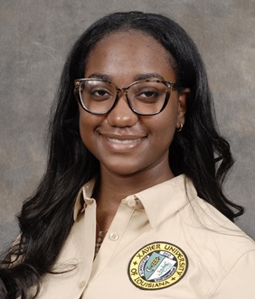 Professional head-and-shoulders portrait of a person with long, dark wavy hair wearing eyeglasses and a light tan collared polo shirt. A circular Xavier University of Louisiana logo is visible on the left side of the shirt. The person is facing the camera against a neutral gray mottled studio background. The image is framed from the chest up and lit with soft, even studio lighting.