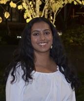 Outdoor portrait of a person with long, dark wavy hair wearing a white blouse. The person is standing and facing the camera in an evening garden setting. Decorative tree lights and foliage are visible in the background, creating a warmly lit backdrop against the darker surroundings.