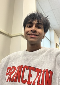 Indoor portrait of a person with short, dark hair wearing a light gray sweatshirt. The person is facing the camera in a hallway or classroom setting with beige walls and a drop ceiling visible in the background. The sweatshirt features large red text reading “PRINCETON.” The image is evenly lit with indoor lighting.